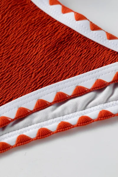 Macro close-up of textured orange bikini fabric, white edge binding, orange zigzag stitching, and white inner lining.