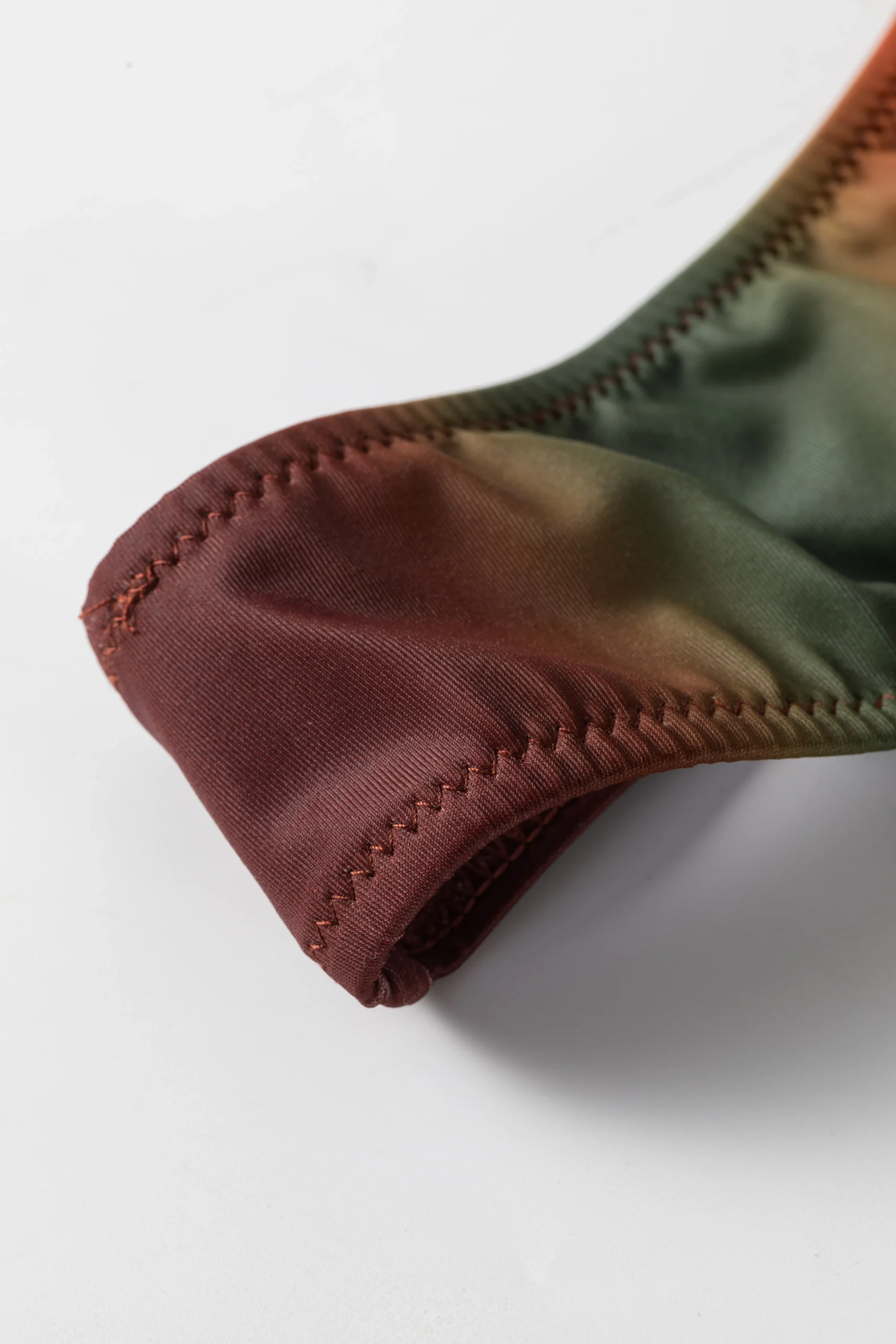 Macro close-up of a fabric edge on the ombre bikini showing zigzag stitching and the transition between rust and olive colors.