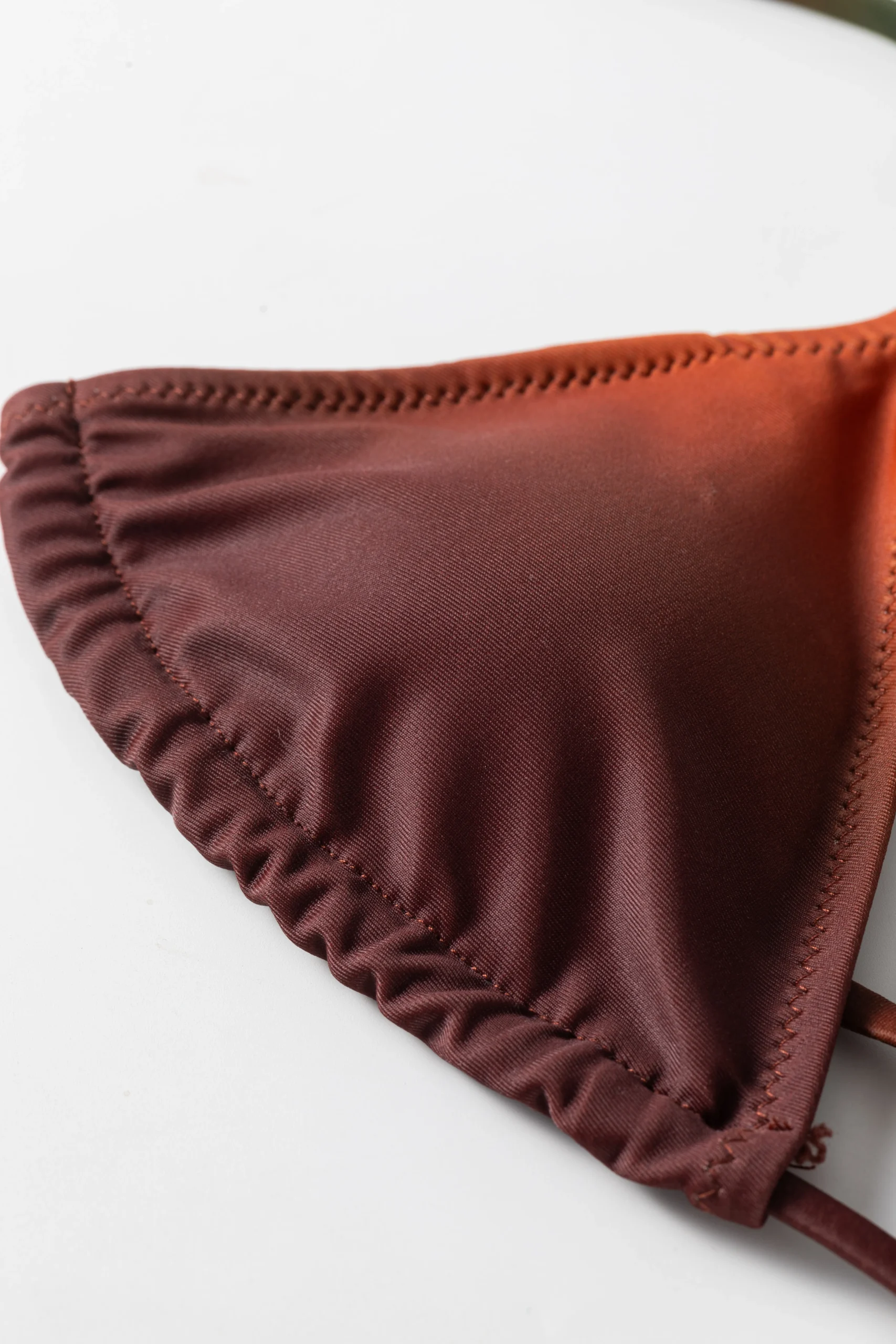 Macro close-up of a bikini top cup edge featuring gathered fabric, zigzag stitching, and a rust-to-orange color fade.