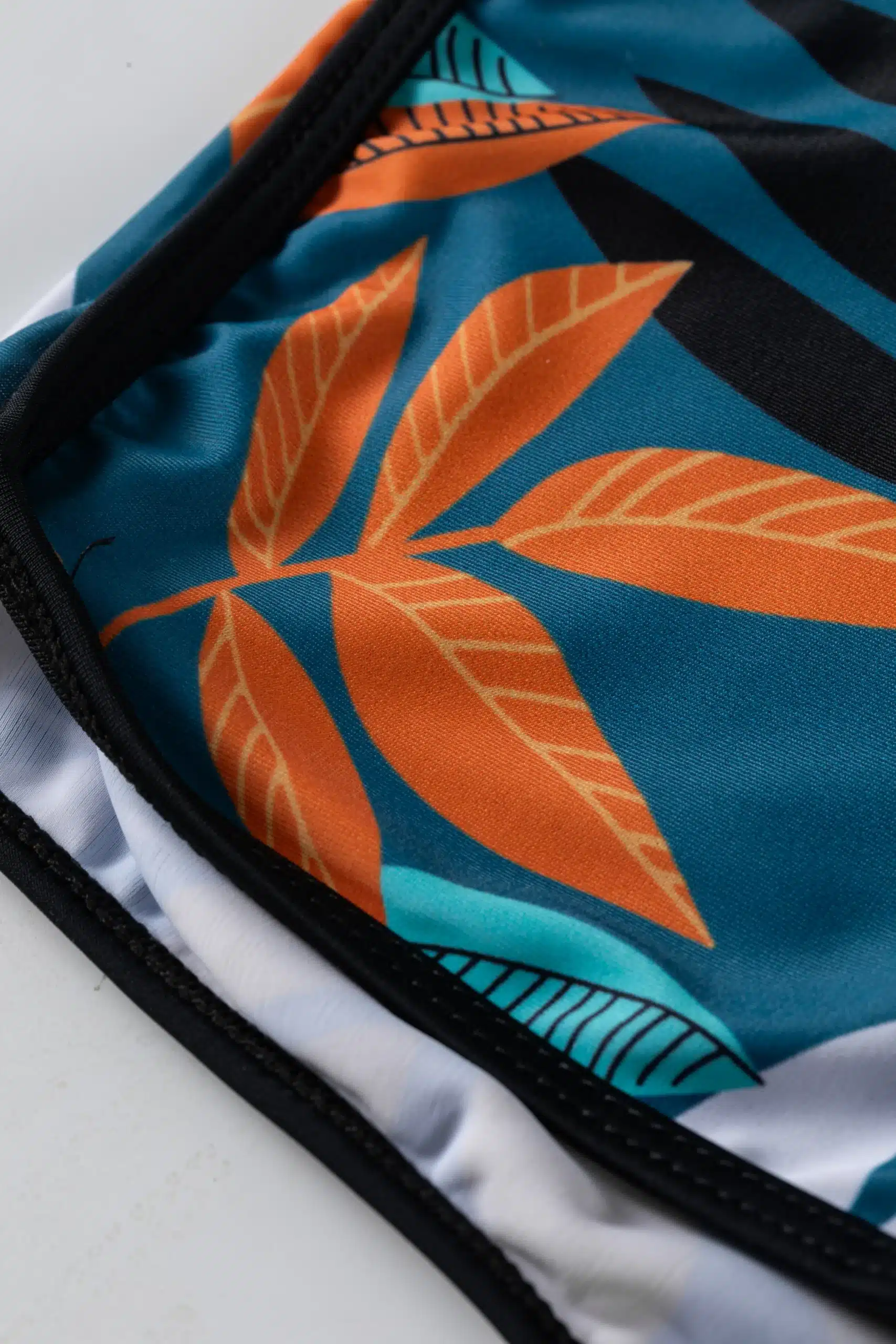 Front flat-lay of a black halter bikini top with a deep V-neck and tie back, paired with teal and orange botanical print swim shorts and a front drawstring.