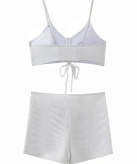 A front flat-lay product shot of a solid white two-piece swimsuit. The top features a slight halter neck with thin tie strings and is bordered with ruffle details along both the top and bottom edges. The matching high-cut bottoms feature side-tie strings and a ruffle trim along the waistband.