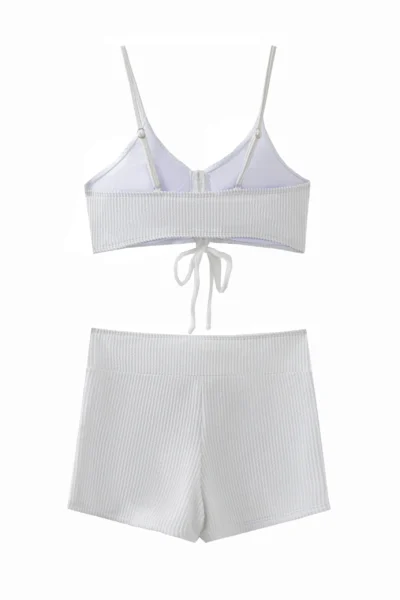 A front flat-lay product shot of a solid white two-piece swimsuit. The top features a slight halter neck with thin tie strings and is bordered with ruffle details along both the top and bottom edges. The matching high-cut bottoms feature side-tie strings and a ruffle trim along the waistband.