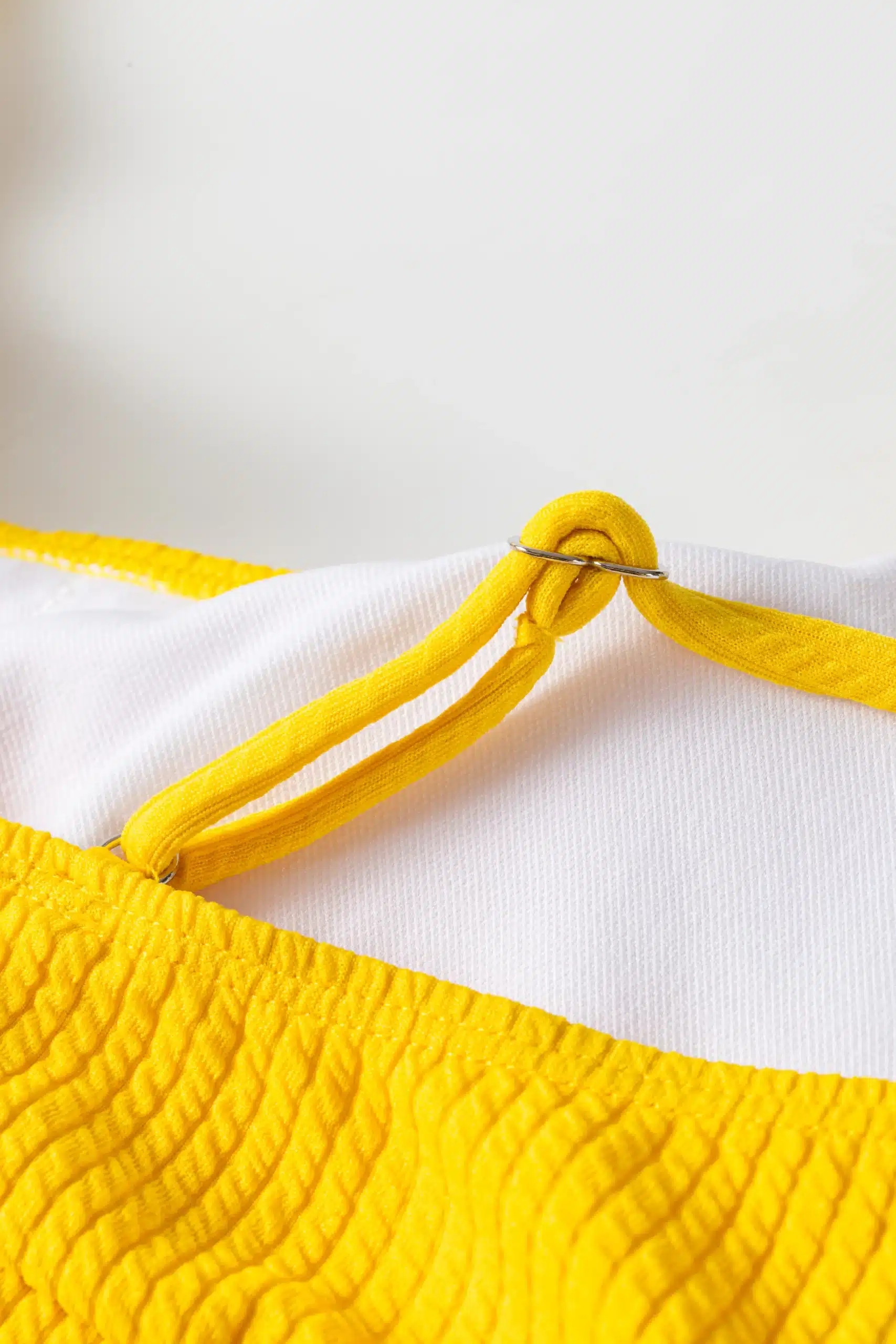 Detail of thin yellow bikini strap looping through a silver metal ring attached to the white-lined top.